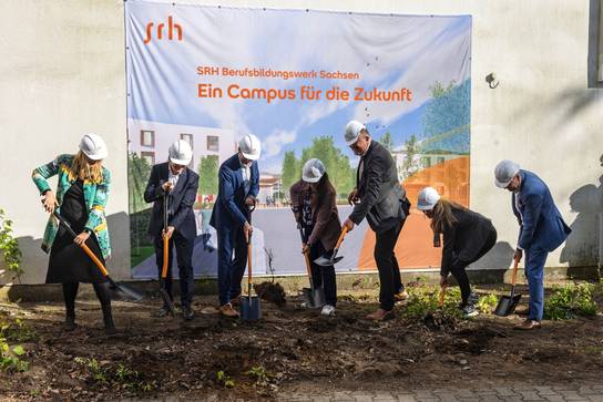 Sieben Menschen mit Helmen auf dem Kopf stechen mit einem Spaten in die Erde zum Start der Arbeiten für den neuen Campus. 